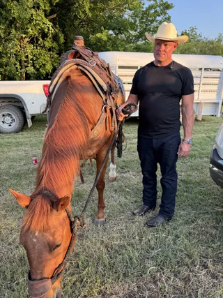 Shawn stands outdoors wearing a cowboy hat and black t-shirt while holding a saddled brown horse by its lead rope.